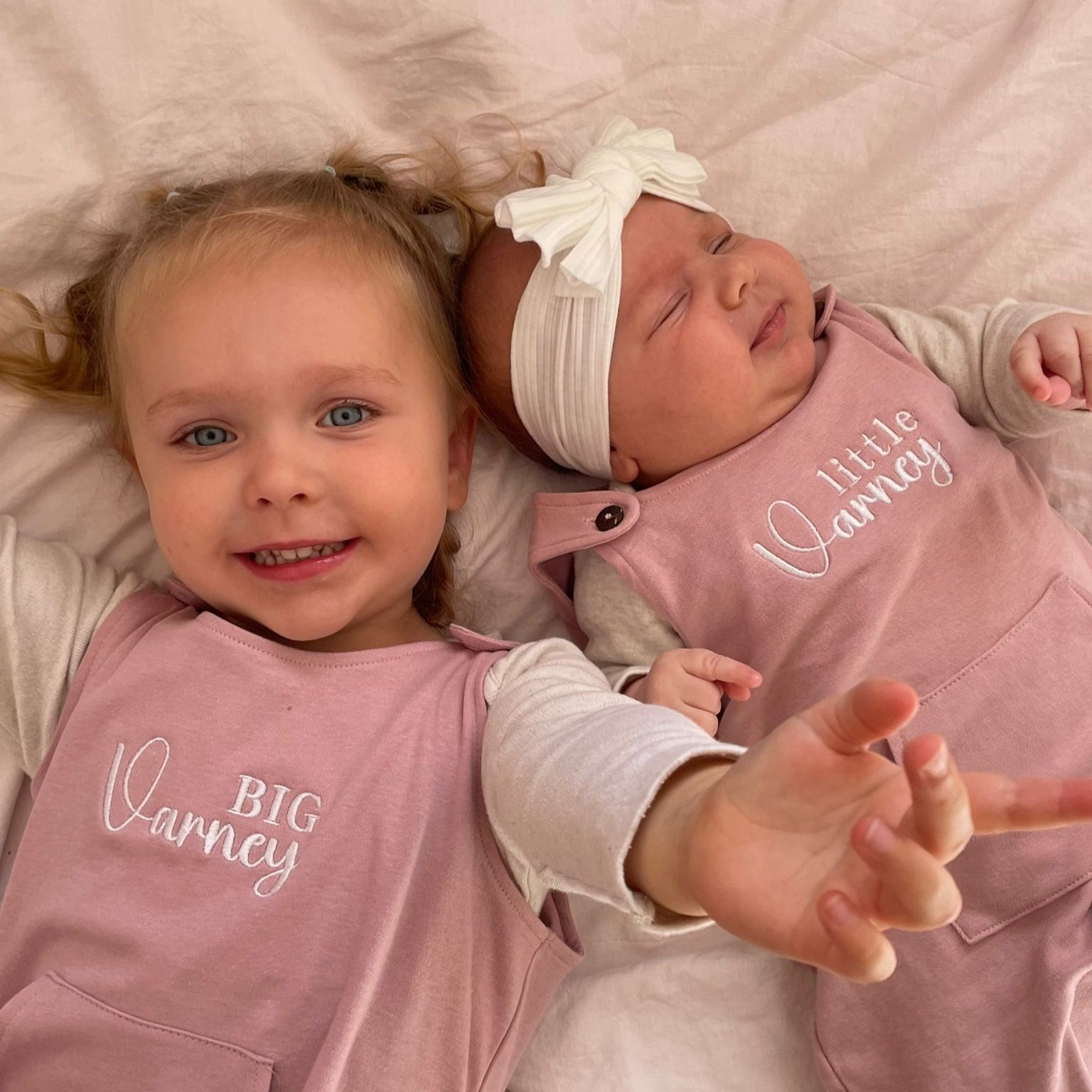 Two young children lie side by side on a light pink blanket, both wearing matching dusty pink dungarees. The older child, smiling brightly, has "BIG Varney" embroidered on her outfit, while the younger baby, wearing a white headband with a bow, has "little Varney" on hers. Both dungarees feature a central kangaroo pocket and button fastenings at the shoulders.